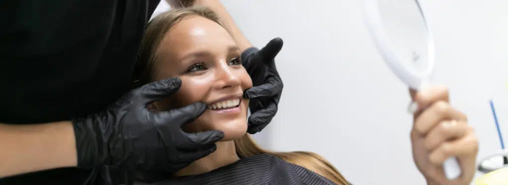 Smiling girl looks in mirror while cosmetic dentist points to her teeth.